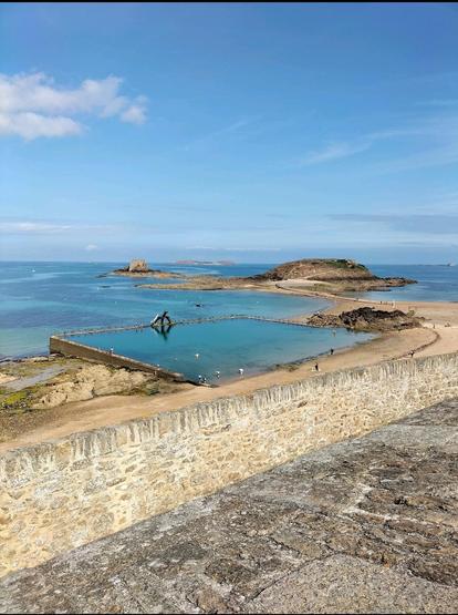 Vue depuis les remparts sur une grande piscine naturelle en bord de mer, entourée de sable et de rochers. Au loin, une île accessible à marée basse, et une mer turquoise sous un ciel bleu clair. Quelques personnes se baignent dans le bassin rectangulaire