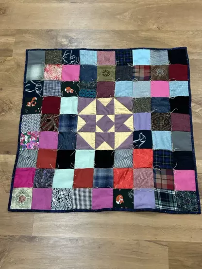 A baby quilt made up of lots of squares tied in the corner. The center block is a kind of star pattern in a purple corduroy and cream silk background.