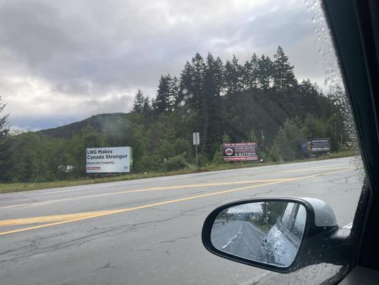 A picture from out a car window shows two large billboards alongside a highway. There are trees behind. The sky is cloudy. The billboard of interest is white with black text that says “LNG MAKES CANADA STRONGER — LNGFACTS.CA”