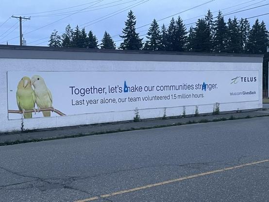 A large and long Telus billboard features their cute-animal inspired yellow budgies on the far left of the billboard and the TELUS logo on the right. In between in two long lines of text it says
“Together, let’s make our communities stronger. Last year alone, our team volunteered 1.5 million hours.”

The “m” in make and “o” in stronger have been spray painted over with blue paint with a “b” and “a” respectively spelling “bake” and “stranger”.