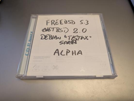 A CD jewel case labeled by hand with several operating system names and versions: "FreeBSD 5.3", "NetBSD 2.0", "Debian 'Testing' Sarge", and "ALPHA". The case insert contains 3 TDK CD-R Speed-X discs. The handwriting suggests the CD contains installation or boot media for Alpha architecture systems.