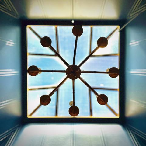 A square skylight is viewed from directly below its central, aligned chandelier hanging beneath, as bright blues and metallic bronzes reflect off its four wooden walls.