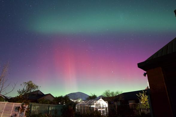 In the foreground is our brick home, beyond that is our backyard with a vegetable garden and our green house. In the far background is Saddle Hill in Mosgiel. In the sky above is the aurora australis, at the bottom is shades of light green, above that are shades of red and purple with rays of white. Above those colours is a band of teal green in the sky. There are stars scattered throughout the sky.