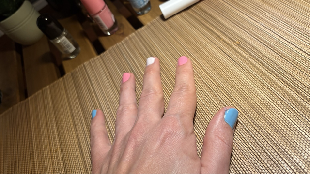 My hand with painted nails, a finger each light blue, light pink, white, pink, blue, to form a trans flag with the hand. Some nail polish bottles still in the background.