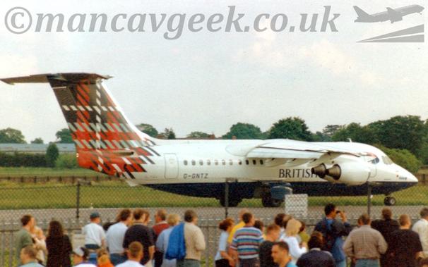 Side view of a high-winged, 4 engined jet airliner taxiing from left to right along a grey runway.
The plane is mostly white, with a blue belly, and "British" titles on the lower forward fuselage.
The black registration "G-GNTZ" is on the lower rear fuselage, while the tail and rear fuselage is covered in a black, grey, red, and white tartan.
In the foreground, a large group of people is gathered around, some looking at this plane but mostly staring in various, different directions, with a black mesh fence between them and the runway.
Green grass in the background leads up to trees in the distance, under cloudy white sky.