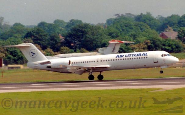 Side view of a white, twin engined jet airliner with the engines mounted on the sides of the rear fuselage, flying from left to right at a very low altitude, maybe a metre above as runway, with the undercarriage lowered, flaps extended from the rear of the wings, and the nose fairly flat, suggesting it is about to land.
Green grass fills the foreground, lining the side of the runway, with more grass in the background, leading up to trees in the background.
The rather dilapidated white fuselage of a 3 engined jet airliner is visible in front of the trees, facing to the left.
Grey sky fills the rest of the frame.