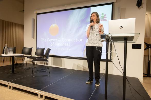 Sarah, a woman with light skin tone and shoulder length light brown hair, standing on a stage holding a microphone. To one side of her is a laptop on a stand. To the other are several empty chairs around a small table. Behind her is a screen showing a slide with text on it reading “the power and connection of kindness.”