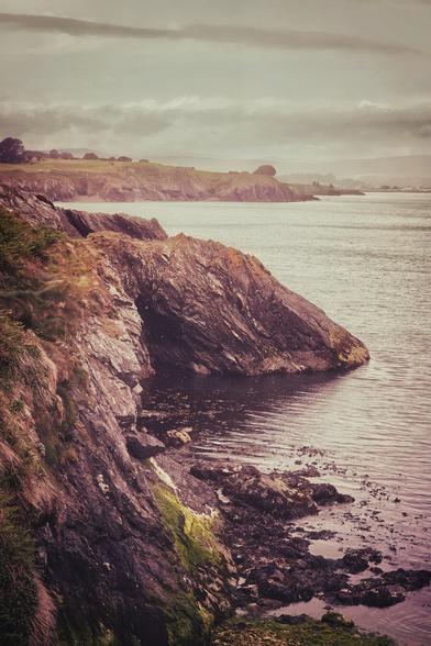 A dramatic coastal scene featuring a rugged, rocky cliff face meeting the calm waters of the sea under a cloudy sky. The cliffs show various textures and colours, with some green moss clinging to the darker rock. In the distance, the Wicklow coastline stretches further, with a headland and hints of a town or village on the horizon. The overall mood is slightly muted and contemplative, with a vintage aesthetic.