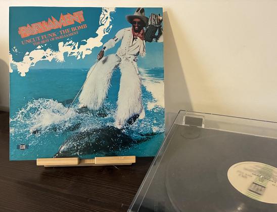 vinyl record cover of "Uncut Funk – The Bomb: The Best of Parliament”, displayed next to a turntable playing the record. The cover displays a man dressed in white with furry pants, holding a boom box on his shoulder, skiing over the sea on two dolphins.