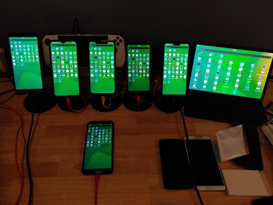 A bunch of postmarketOS devices all running Phosh with default postmarketOS wallpaper and the same set of apps installed.  From left to right:

* PinePhone Pro
* OnePlus 6T
* Xiaomi Poco F1
* Google Pixel 3a
* OnePlus 6
* Xiaomi Pad 5 Pro
* PinePhone (in the front of the other devices)