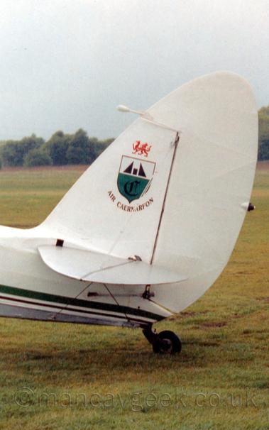 Close-up of the tail of a World War 2-era passenger plane parked on grass facing to the left.
The plane is mostly white, with  a green and grey stripe running along the body, 
There is a green and white shield logo on the white tail, above black "Air Caernarfon" titles, and below an image of a red dragon facing to the left.
In the background, grass leads up to trees in the distance, under grey sky.