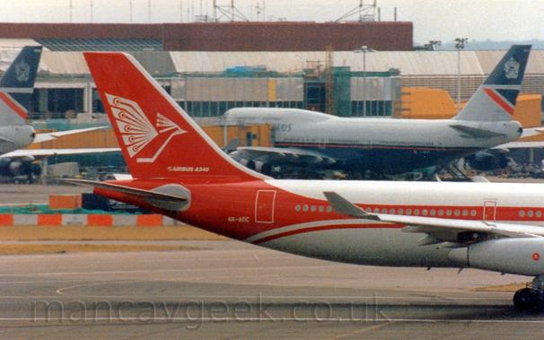 Side view of the rear fuselage and tail of a 4 engined jet airliner moving from right to left at a high speed.
The plane is mostly white, with a thick red stripe running along the body, sweeping up into the tail and around the lower rear fuselage.
There is a stylised white flying bird on the tail, above white "Airbus A340" titles, and the white registration "4R-ADC" on the lower rear fuselage.
In the background, a grey terminal building stretches across the frame, with bright yellow airbridges attached to  a very large, grey and dark blue jet airliner, with the tail of a similar plane coming in to view on the left of the frame.