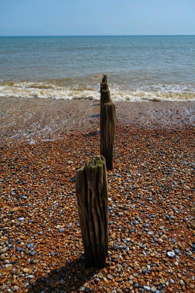 A vertical, high-angle shot captures a shingle beach receding towards the sea under a clear blue sky. In the foreground, four weathered wooden groynes stand upright in a line, stretching from the shore into the gently lapping waves. The beach is a mix of pebbles and stones in shades of brown, grey, and ochre. Small waves with white foam break softly on the shingle, and the sea beyond appears calm.