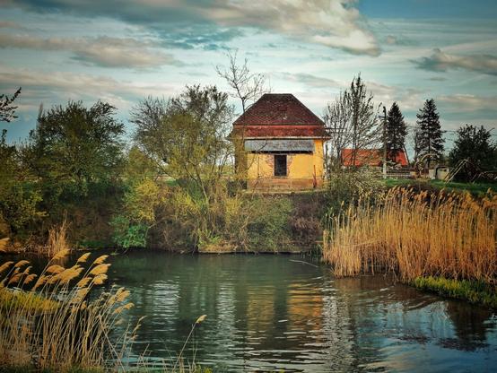 The image shows a small, weathered, yellow-orange building with a reddish-brown tiled roof, situated on the far bank of a body of water. The building has a dark, rectangular opening, which used to be a window, with a sloped roof addition over it. The building is surrounded by various trees and bushes, some with light green foliage, indicating spring. 
In the foreground, there is a body of water with reflections of the sky and the trees. The water's edge in the foreground and on the right bank is lined with tall, dry, light brown reeds. The sky above is partly cloudy, with areas of blue and patches of white and grey clouds. The overall impression is that of a rural or natural setting.