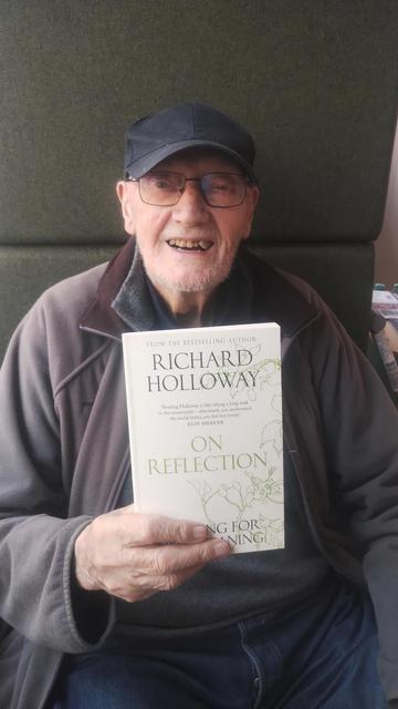 Reverend Richard Holloway smiling and holding up a copy of his latest book, On Reflection, in the Edinburgh Bookshop.