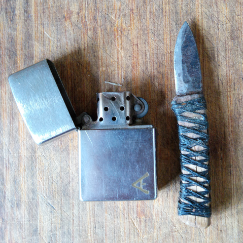 Ein kleines geschmiedetes Messer liegt neben einem Zippofeuerzeug. Es hat einen mit Garn umwickelten Holzgriff.