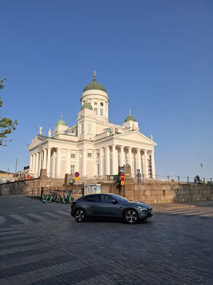 A white, Neoclassical church stands on a platform. The square church has triangular tympana supported by Corinthian pillars on the two visible sides and sports a tall green dome in the center, with smaller green domes on the corners. The domes are all topped with golden Latin crosses. Statues of the Apostles are placed on top of the tympana.