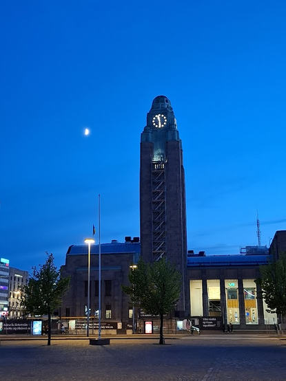 A monumental clock tower shows that it is half past eleven. The half moon is visible in the cerulean sky.