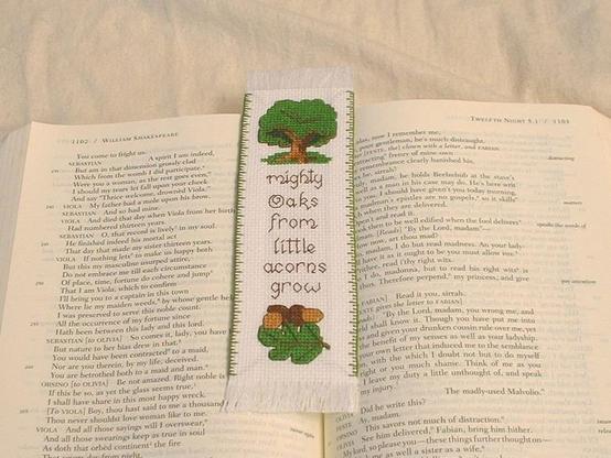 a cross stitched bookmark laid out on top of an open book. the pattern is of a bushy tree above and an oak leaf and two acorns below the text that reads: Mighty Oaks from Little Acorns Grow.
