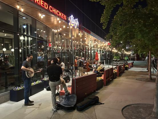 A lively nighttime scene outside a restaurant with a sign for fried chicken. Musicians play instruments, including a banjo, while people gather and enjoy the atmosphere, adorned with string lights and planters.