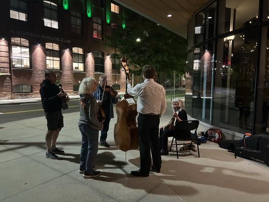 A small group of musicians plays instruments outside the Cambridge Athletic Club at night. The scene features a mix of string instruments, with a couple of musicians standing and others seated. The setting is illuminated by building lights and street lamps, creating a cozy atmosphere.