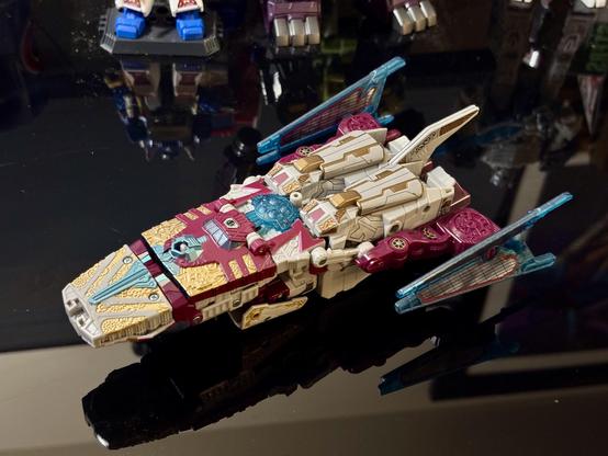 Transformers Legacy Vector Prime with the Toyhax stickers and leg parts applied, placed in spaceship mode on a black table with the feet of other Transformers robots in the background.