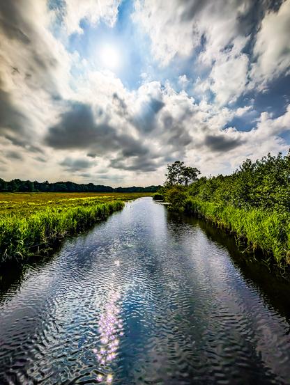 A serene, straight canal flowing under a dynamic sky. The sun, partially obscured by clouds, casts a bright spot in the upper center, with rays of light radiating downwards. These rays create shimmering reflections on the surface of the water. The canal itself acts as a strong leading line, guiding the viewer's eye towards the horizon. On both sides, vibrant green vegetation lines the banks.