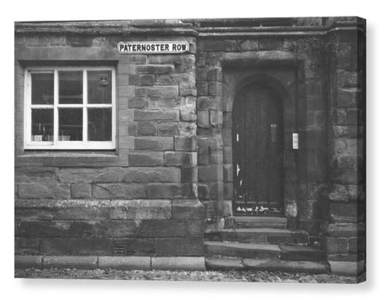 Vintage looking black and white photograph of an arched wooden door within an old stone surround. Above there is a street sign, Paternoster Row.  The image is shown printed upon a box canvas.