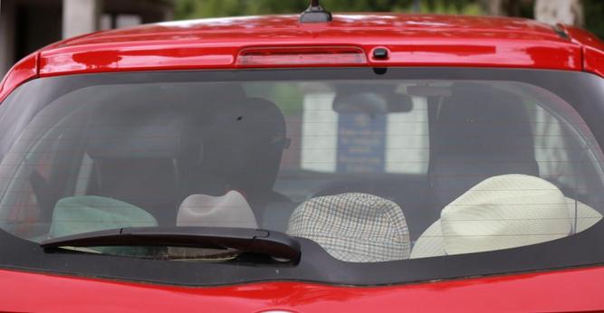 The rear window of a red car, a close crop. Four hats of various sorts including a Trilby or Fedora, a checked hat possibly of a deerstalking nature, and a Panama are resting across the parcel shelf.