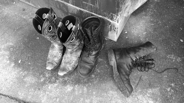 A black and white photo of 4 boots. 2 pair. 1 pair of cowboy boots and 1 pair of combat style boots. One of the combat boots is tipped over.