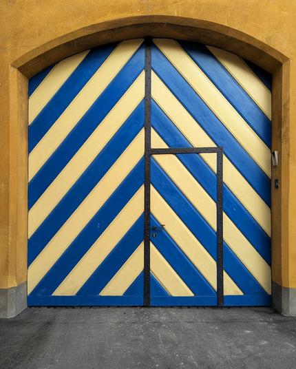 A large, arched wooden gate featuring diagonal stripes of blue and yellow. A smaller pedestrian door is set into the lower right of the gate, also striped and framed in black. The gate is set into a yellow-ochre wall, with a grey paved ground in front.