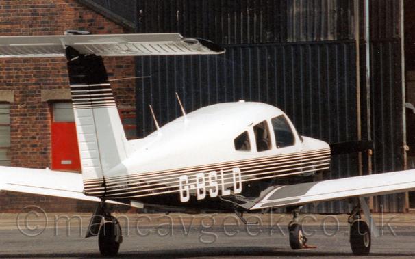Rear side view of a single-engined light aircraft parked facing to the right and away from the camera.
The plane is mostly white, with a black belly, and thin black and white stripes along the middle, overlaid with the white registration "G-BSLD" on the lower rear fuselage.
The colours are inverted on the tail, black at the top and white at the bottom.
The plane is parked on black tarmac, facing a brown brick hangar with large black metal doors.