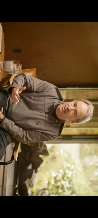 The screencap shows Linus Torvalds sitting at a table in a warm, indoor setting with large windows that allow natural light to fill the room. He is wearing a gray hoodie and has gray hair. The man is gesturing with his hands, suggesting he is engaged in a conversation or explaining git. The table has a glass container, possibly a water bottle, and a brown leather chair is visible. The background features a blurred view of greenery outside, indicating a peaceful environment. The overall atmosphere is calm and relaxed.