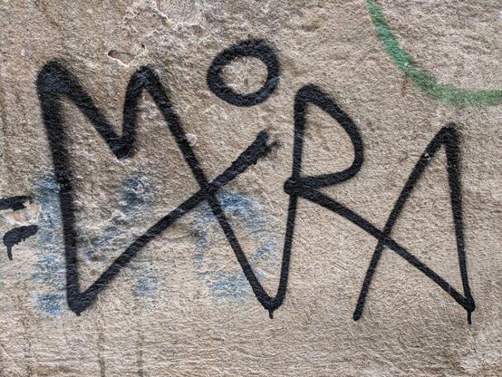 A close-up of a weathered, light brown wall with textured plaster. The word "MIRA" is spray-painted in thick, black, stylised capital letters across the centre. The 'I' has a circle above it, and the 'M' and 'R' have a more angular, graffiti-like appearance. Faint smudges of blue and green paint are visible underneath and to the side of the black lettering.