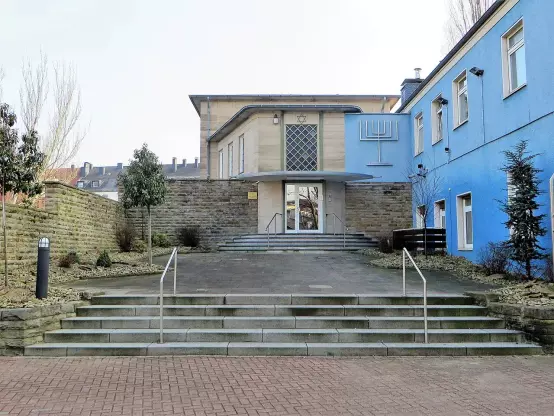 The image depicts a building with a modern design, featuring an entrance with a set of stairs leading up to it. The building has a combination of stone and blue-painted walls. Surrounding the entrance, there are some plants and landscaping elements, and the overall setting appears to be urban or suburban. There are no people visible in the image.

Image Credits: Wikimedia / Bärwinkel,Klaus / CC BY 3.0