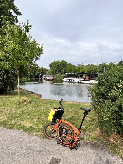 Un vélo pliant Brompton devant une étendue d’eau où se trouve une péniche, entouré de verdure. Il y a un rayon de soleil