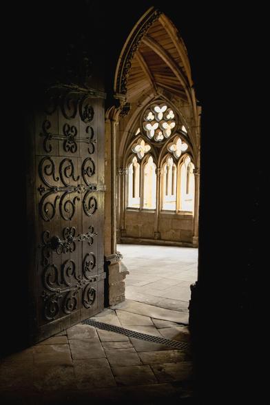 Inside a gothic cathedral.  The arched, wooden door is open offering a view towards the cloisters.