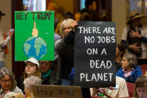A woman at a protest, holding up a sign that reads, "There are no jobs on dead planet." Next to her is a sign that reads, "Hey Trump," and the Planet Earth is giving him the middle finger.