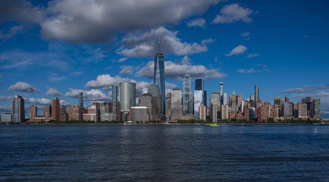 A view of Downtown Manhattan as seen from Downtown Jersey City.  There are fluffy clouds and a very blue sky, and the colors of the buildings are vibrant.  A fluorescent green speedboat is on the Hudson River proceeding from the viewer's right to left.