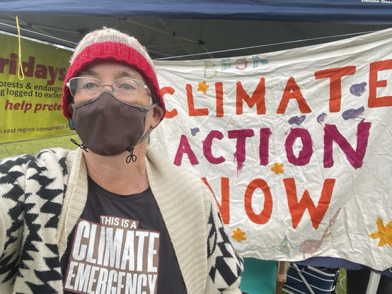 Masked woman in beanie, mask and Climate Emergency tshirt stands in front of a Climate Action Now