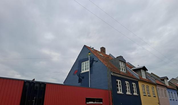 Short version: a Mary Poppins mural painted around a window three-storeys up on a gable wall.

Long full description: We’re standing on the corner of Ole Rømers Gade og Montanagade in Frederiksbjerg, mid-city Aarhus, Denmark – a lovely mixed-used area with lovely narrow residential streets. Looking up and beyond, we see the top storey of a row of centuries-old picturesque two-storey houses. Each has a sharply sloping red-tiled triangular roof. Each is painted a different elegant pastel colour – dark blue, yellow, pink, light blue. The windows are multi-paned and white. Each has one dormer attic window. On the corner, a high, modern, designer wall of deep red vertical wooden planks with a black striped section for a door. There is a narrow elongated slit of a window in it. Rising beyond the fence: the deep blue gable wall of the corner house. It has a red-brick chimney and a single multi-pane white window on the top floor. Painted up the full three storeys: a Mary Poppins mural. The chimney sweep is a silhouette swinging off the top of a painted white lamppost right beside the window. It rises the full length of the gable wall, topped off just under the roof with a glowing white lantern. Floating up off to the right, a little lower down: a silhouette of Mary Poppins holding her red umbrella open above her. Above them, high electricity wires criss-cross below a metal-grey rain-promising cloudy sky that Mary Poppins seems about to float off into.