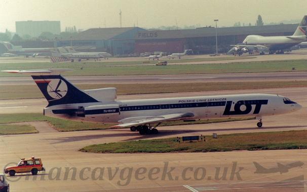 Side view of a 3 engined jet airliner with the engines mounted on the rear fuselage, taxiing from left to right.
The plane is mostly white, with a thick blue stripe running along the body, covering the passenger cabin windows, as well as  over the engines mounted on the sides of the fuselage.
There are also large, blue "LOT" titles on the forward fuselage, with smaller blue Polish titles that translate into English as "Polish Air Lines".
The tail is the same dark blue, with a white circle containing a stylised image of a blue flying bird.
Grey concrete apron fills the foreground, with green grass bordering taxiways and a runway in the background leading up to a red and grey hangar next to a blue and grey hangar, both with planes parked in front, under bright but hazy sky.