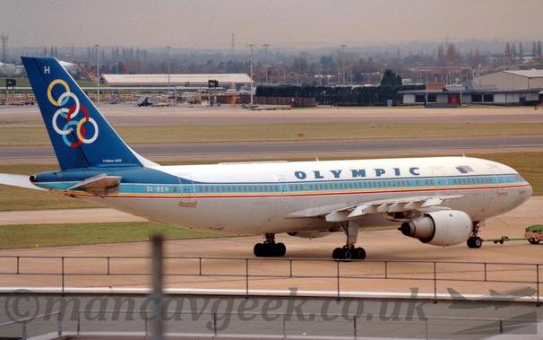 Side view of a twin engined jet airliner facing to the right but being pushed back to the left by an almost out-of-frame green tug.
The plane has white upper surfaces and a grey belly, with a light blue stripe running along the body covering the passenger cabin windows, with additional blue pinstripe above, and yellow and red pinstripes below.
There are dark blue "Olympic" titles on the upper fuselage over the wings, with a stylised Greek flag partially covered by an EU flag between the forward and mid-cabin doors.
The tail is dark blue, with a series of linked, multi-coloured rings, and the white letter "H" at the top.
The grey flat roof of a building fills the bottom of the photo, while green grass lines a runway and a taxiway in the background, with trees and buildings leading off in to the distance, slowly vanishing into grey haze.