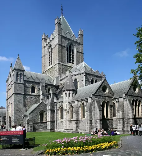 The image shows a large, historic stone church with impressive architectural features, including towers and intricate windows. It appears to have a well-maintained exterior and is surrounded by a garden with colorful flowers. People can be seen sitting on the grass in front of the church, enjoying the pleasant weather. The sky is clear and blue, indicating a sunny day.

Image Credits: Wikimedia / Ingo Mehling / CC BY-SA 4.0