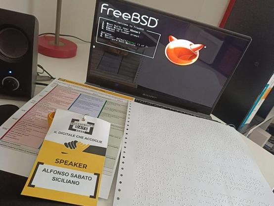 A workspace setup featuring a laptop displaying the FreeBSD boot menu screen. In front of the laptop are several accessibility-related materials: a large braille printout, a color-coded accessibility reference chart, and a yellow conference badge labeled “Accessibility Days 2025 ” with the speaker’s name “Alfonso Sabato Siciliano.”