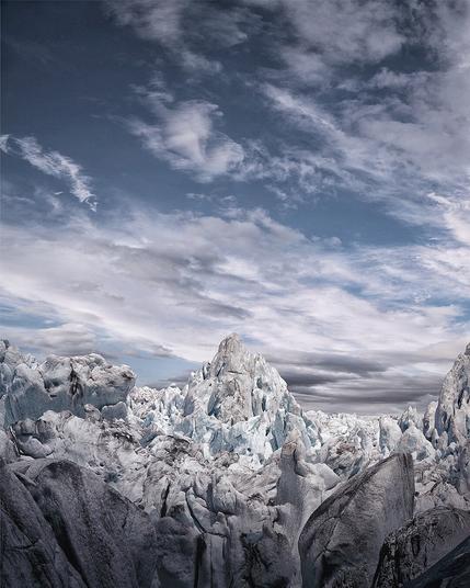 A striking view of a jagged glacier field beneath a dramatic sky. Towering ice formations dominate the scene, their surfaces a blend of frosty blue and weathered grey, suggesting ancient ice shaped by time. The central peak rises sharply like a frozen mountain, surrounded by chaotic ridges and crevices. Above, layered clouds stretch across a blue sky, creating a sense of motion and scale. The overall mood is stark, raw, and awe-inspiring, capturing the harsh beauty of a remote glacial wilderness.