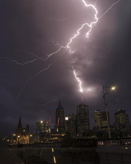 A vivid lightning bolt splits the night sky above Melbourne’s CBD, dramatically illuminating dark storm clouds. The bolt strikes a skyscraper with an orange-lit antenna, casting a sharp contrast against the surrounding buildings lit by office lights. The gothic spire of St. Paul's Cathedral stands prominently on the left, while the bridge in the foreground glistens with raindrops. The city feels alive with tension as nature unleashes its power over the urban landscape.