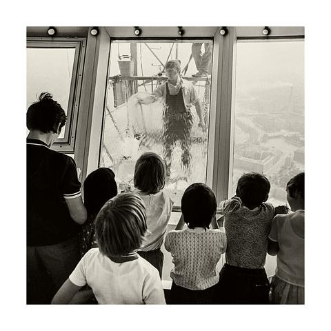 Schwarzweißfoto: Kinder in der Turmkugel des Berliner Fernsehturms, von hinten fotografiert, sehen der Arbeit eines Mannes zu, der die Fenster von außen putzt