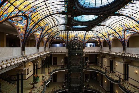 The image depicts an ornate interior space with a grand architectural design. The ceiling is a prominent feature, showcasing a large, arched stained glass skylight with intricate patterns in vibrant colors such as blue, yellow, and red. The skylight is supported by a metal framework, creating a harmonious blend of art and engineering. Below, the space is multi-leveled, with balconies and staircases that add to the grandeur. The walls are adorned with decorative moldings and sconces, enhancing the opulent atmosphere. A central elevator shaft with a dark metal structure is visible, adding to the verticality of the space. The flooring is carpeted, and there are potted plants placed strategically, contributing to the overall aesthetic. The image captures the essence of a historic building, likely a hotel or a public space, with its elaborate design and attention to detail.

Provided by @altbot, generated privately and locally using Ovis2-8B