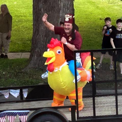 A Shriner in a maroon fez and maroon shirt apparently riding a humongous chicken who is mostly red and orange and yellow but has kind of a rainbowish tail (first four rainbow colors visible: red, orange, yellow, green). (It's a chicken rider costume, of course.) The man is waving and grinning maniacally. They're on a trailer in a parade. (Parade not pictured.)